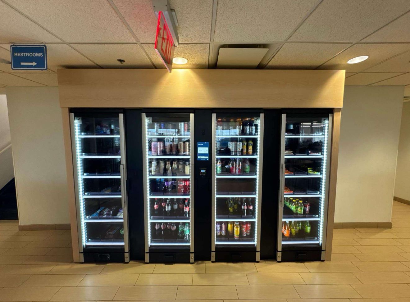 Four illuminated refrigerated vending compartments display beverages in a clean commercial hallway.