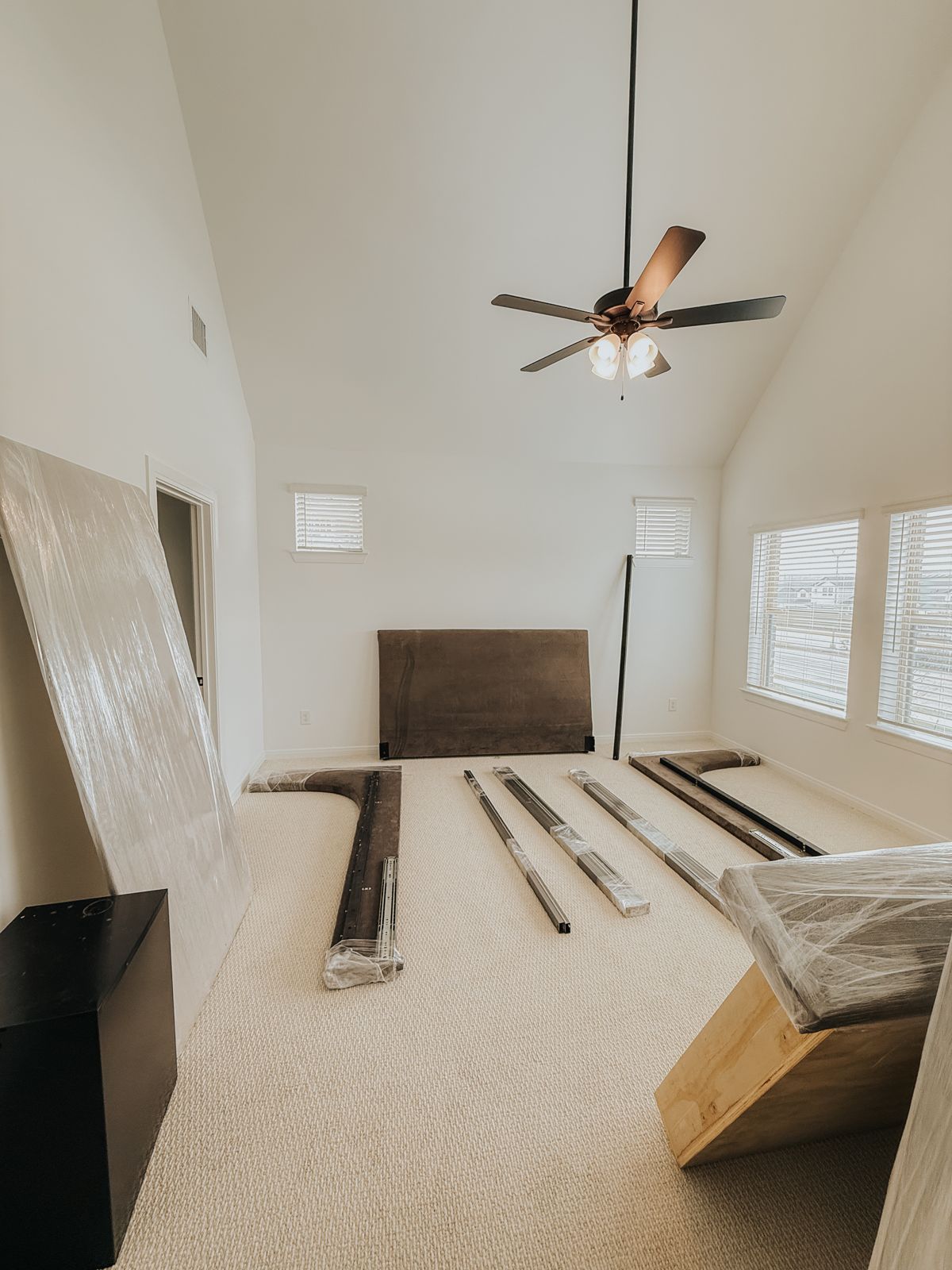 Wrapped bed rails, a headboard, and other furniture components are neatly staged for assembly in a vaulted bedroom.