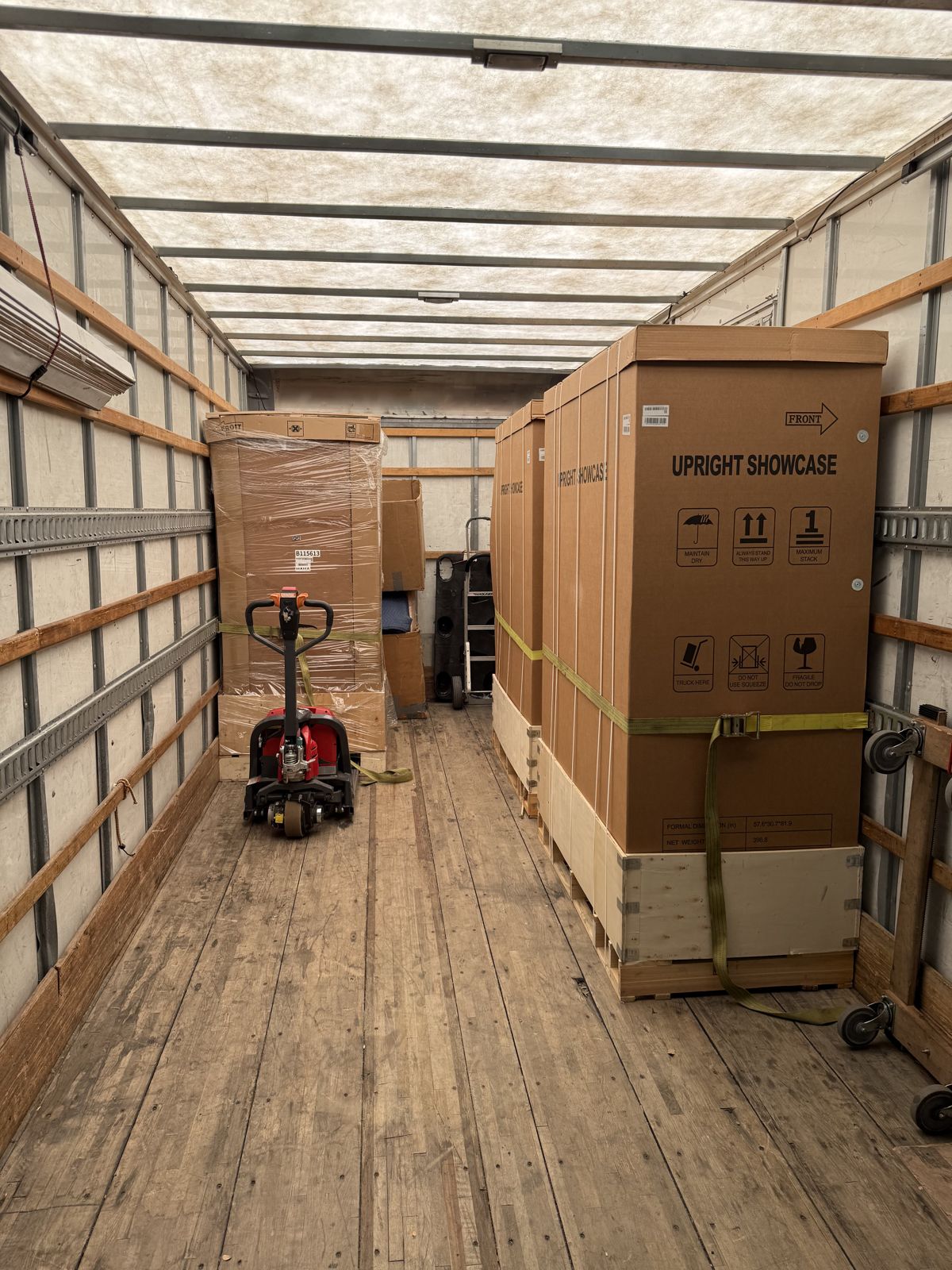 Large boxed upright showcases are strapped securely inside a moving truck beside a powered pallet jack.