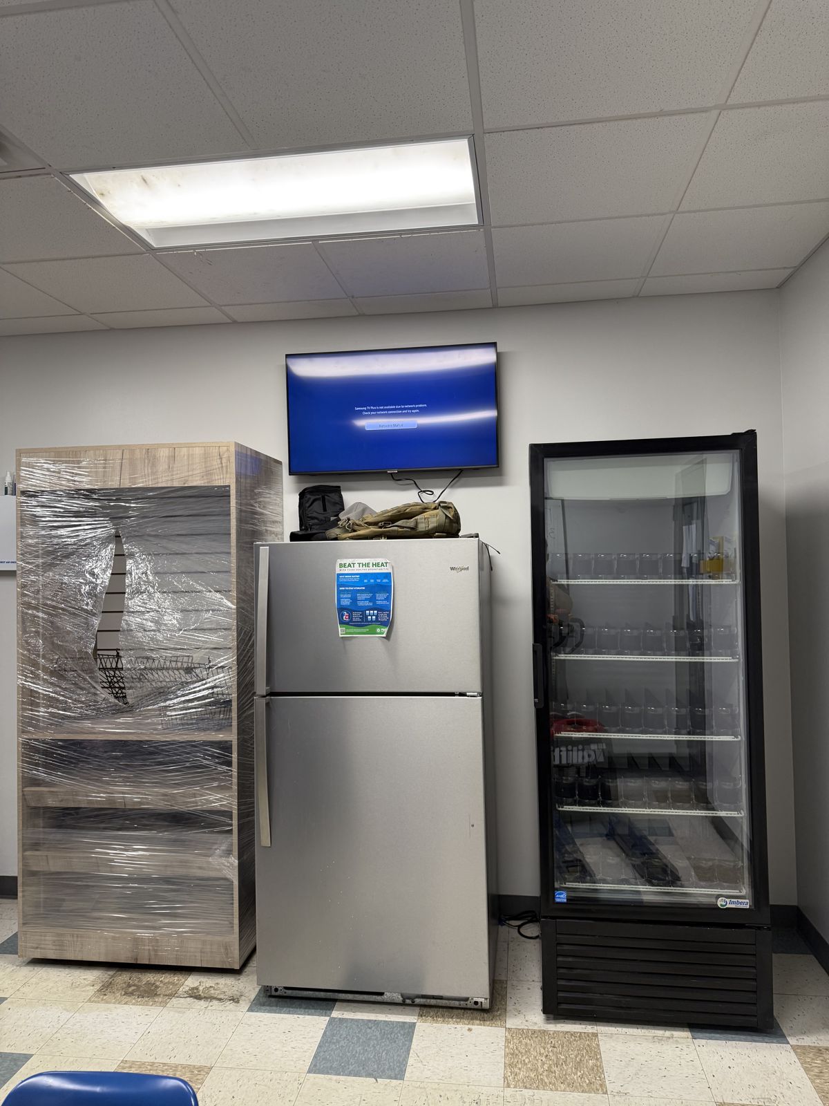 Wrapped commercial equipment, a refrigerator, and a glass-front cooler are positioned inside a breakroom.