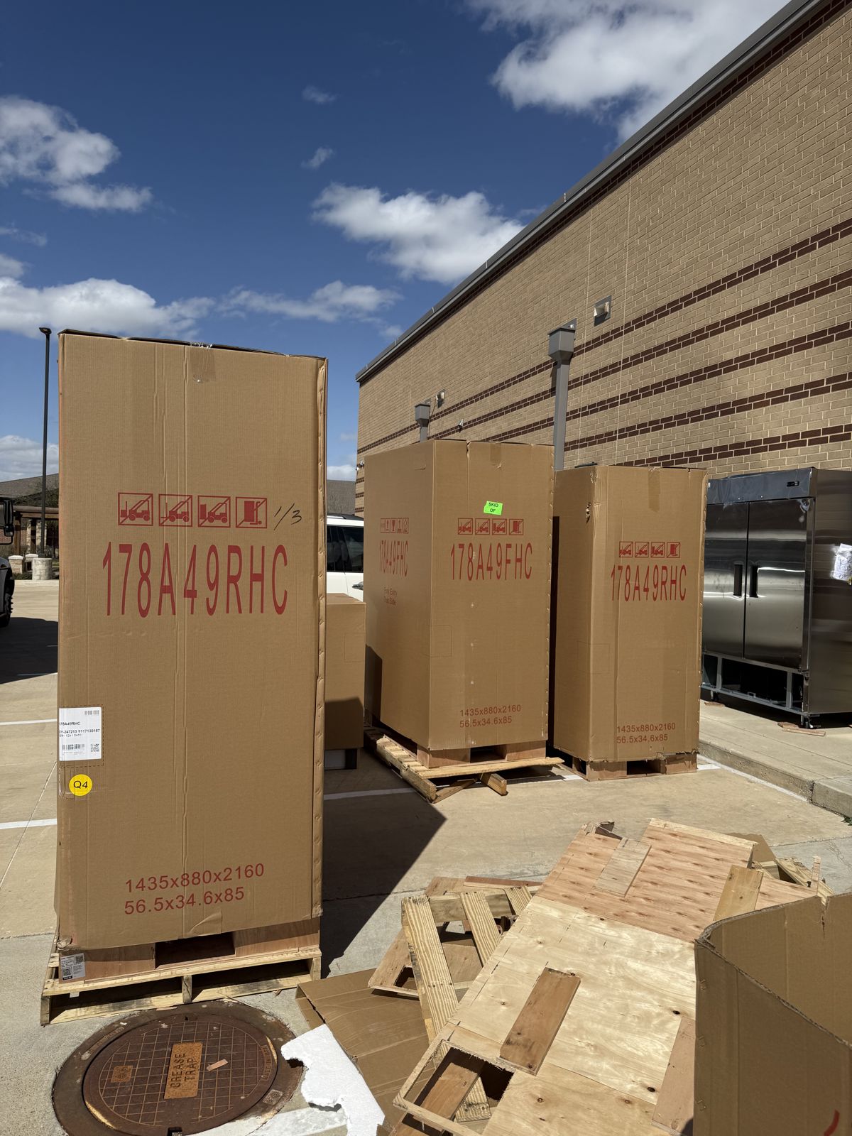 Three oversized commercial items in protective cartons stand on pallets outside a brick building.