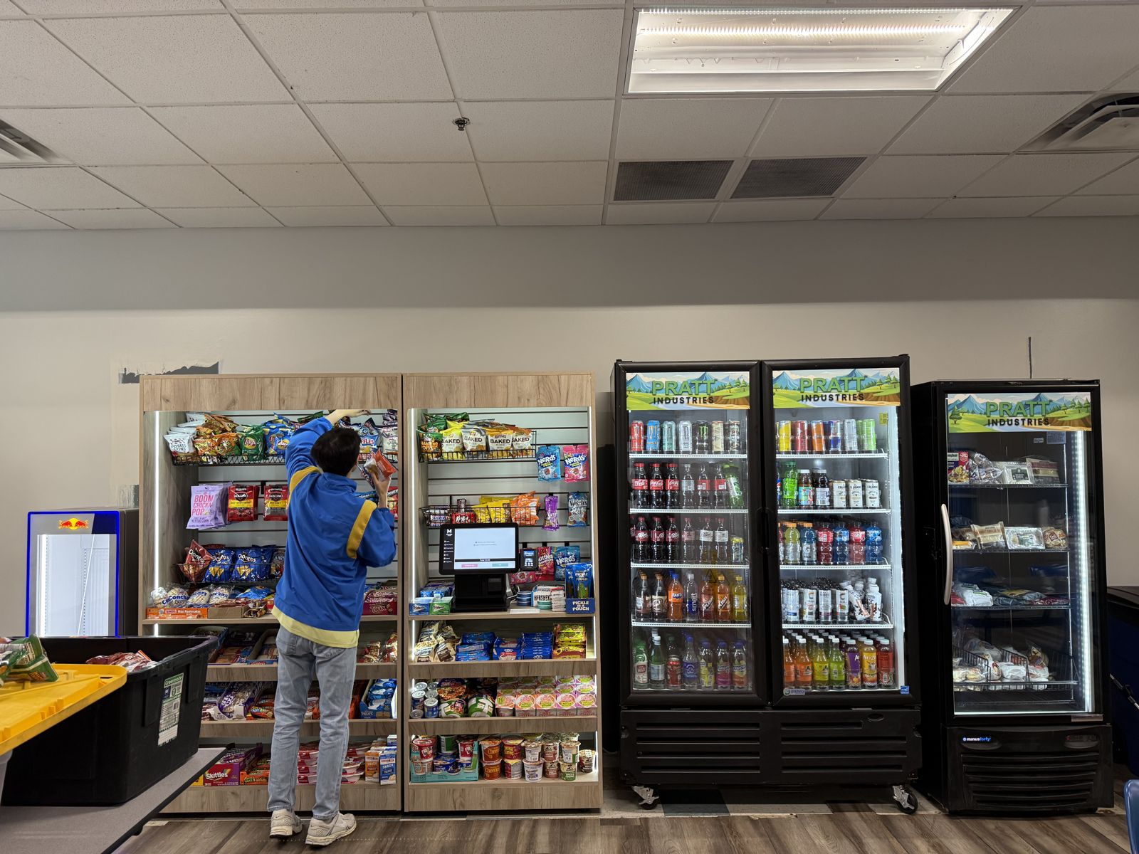 A worker stocks a micro-market display beside several illuminated commercial refrigerators.