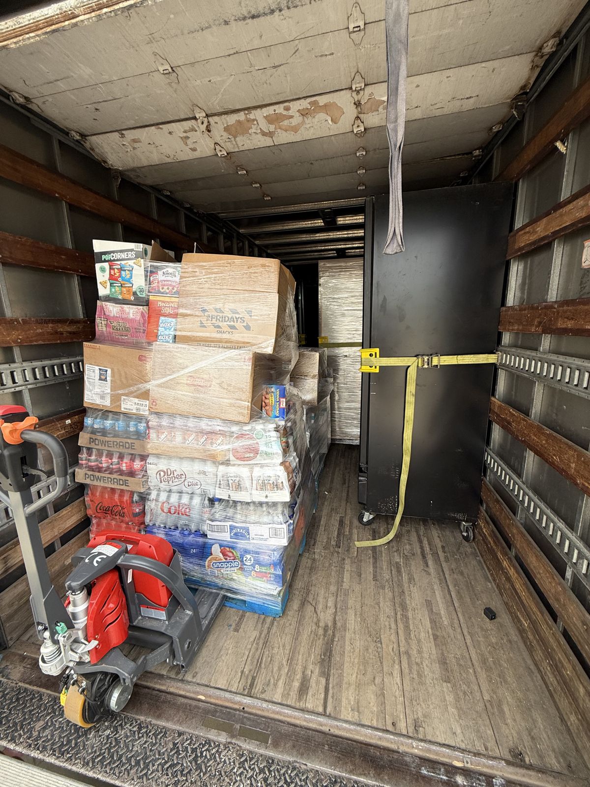 Palletized drinks, boxed snacks, a black cabinet, and a powered pallet jack are secured inside a TL Moving truck.