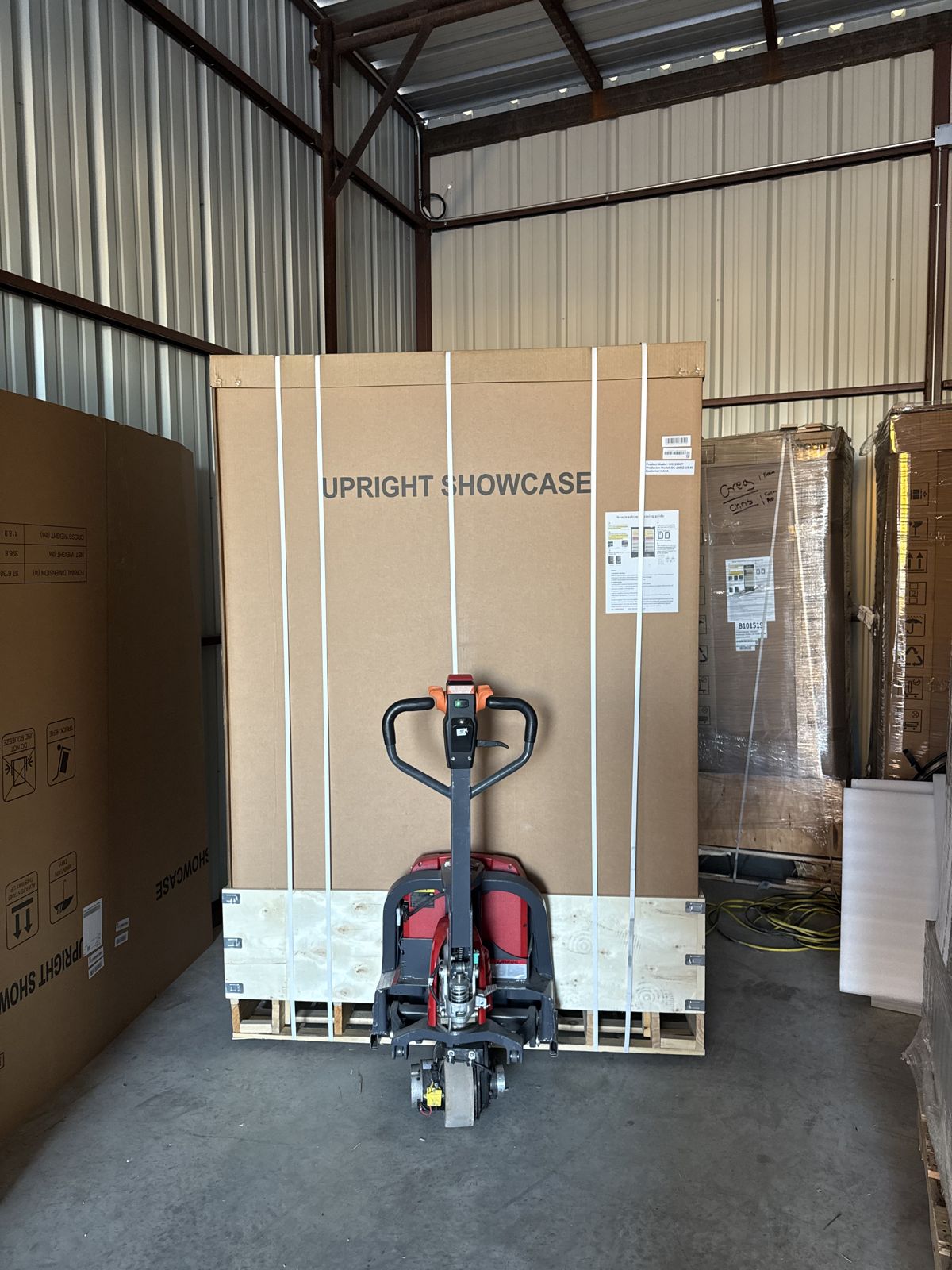 A large crated upright showcase stands on a pallet behind an electric pallet jack in a warehouse.