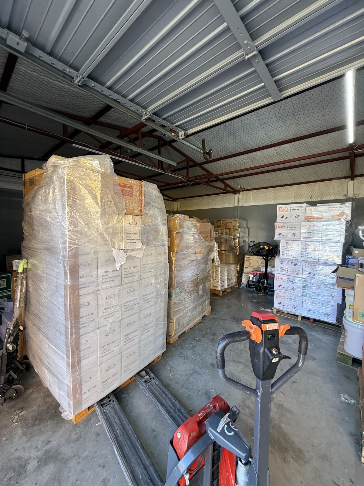 Large shrink-wrapped pallets and stacked cartons fill a warehouse space equipped with pallet jacks.