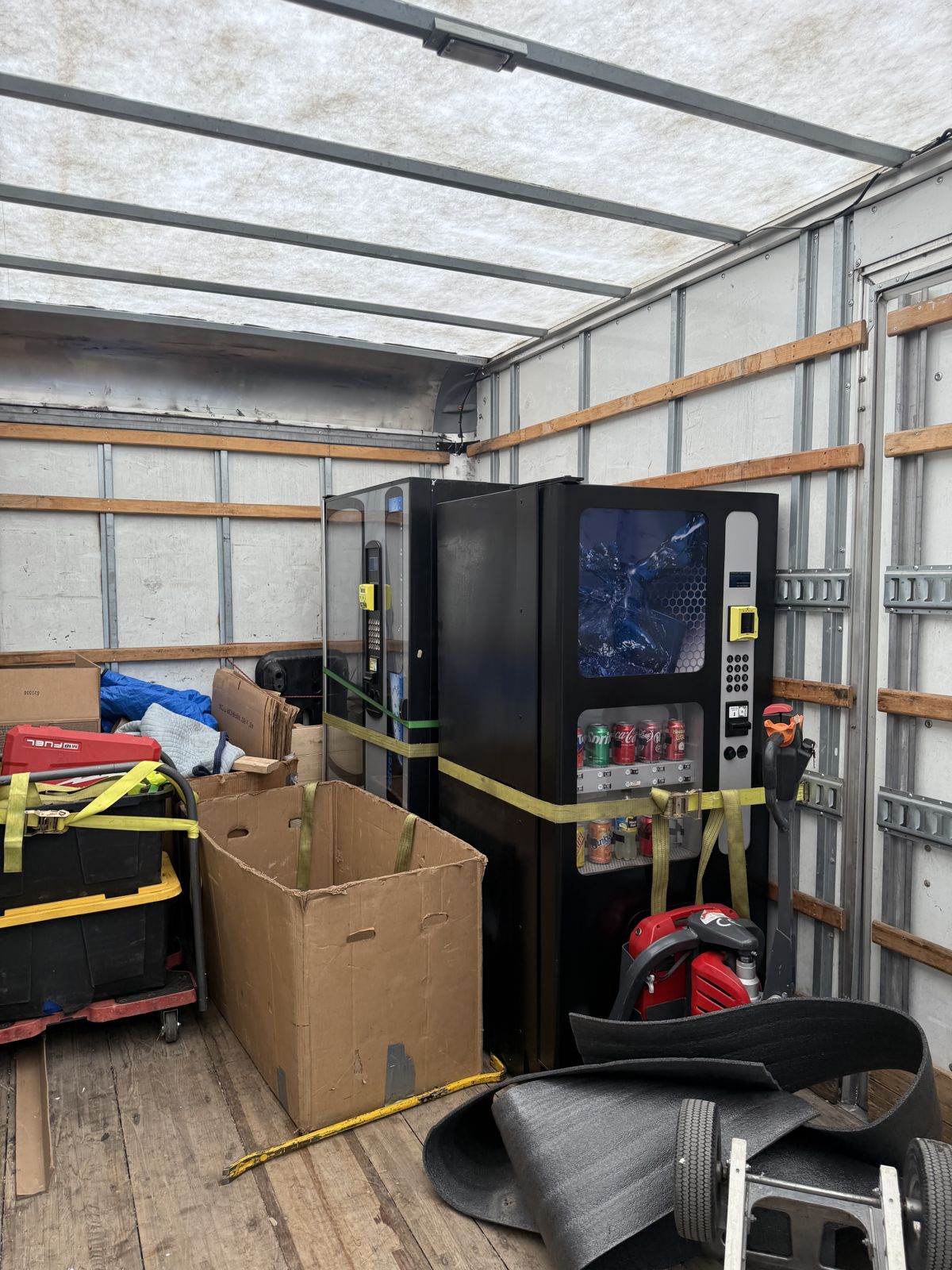 Two vending machines are secured with heavy moving straps inside a box truck alongside moving equipment.
