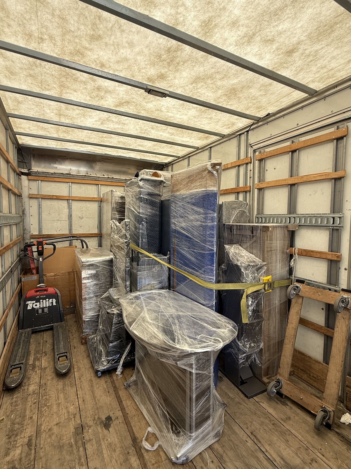 Blanket-wrapped household furniture is secured with straps beside dollies and a pallet jack inside a box truck.