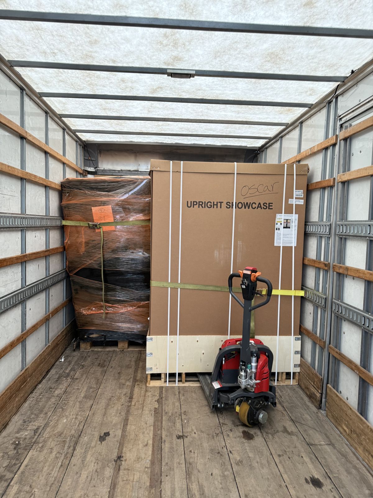 A boxed upright showcase and a wrapped commercial unit are secured beside a powered pallet jack.