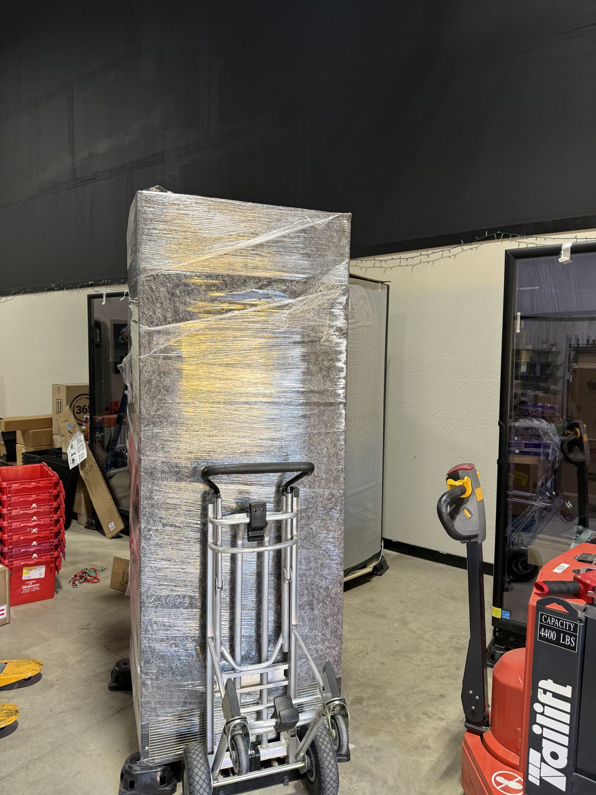 A tall wrapped commercial unit stands beside a hand truck and powered pallet-moving equipment.