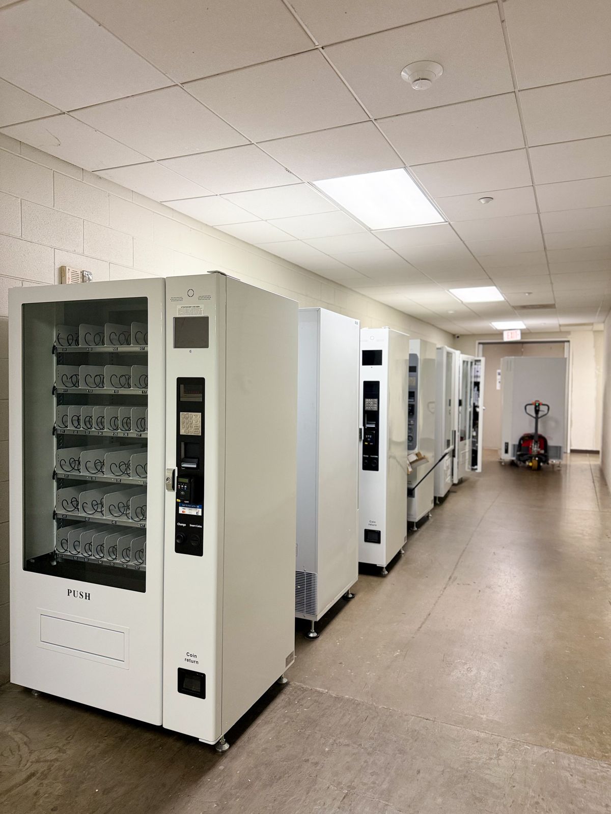 A row of white vending machines lines a bright commercial hallway beside a pallet jack.