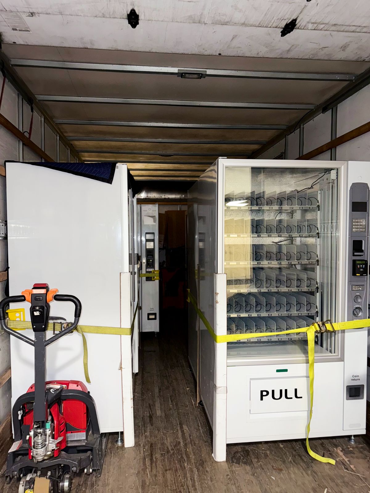 Two vending machines are secured inside a moving truck beside a red electric pallet jack.