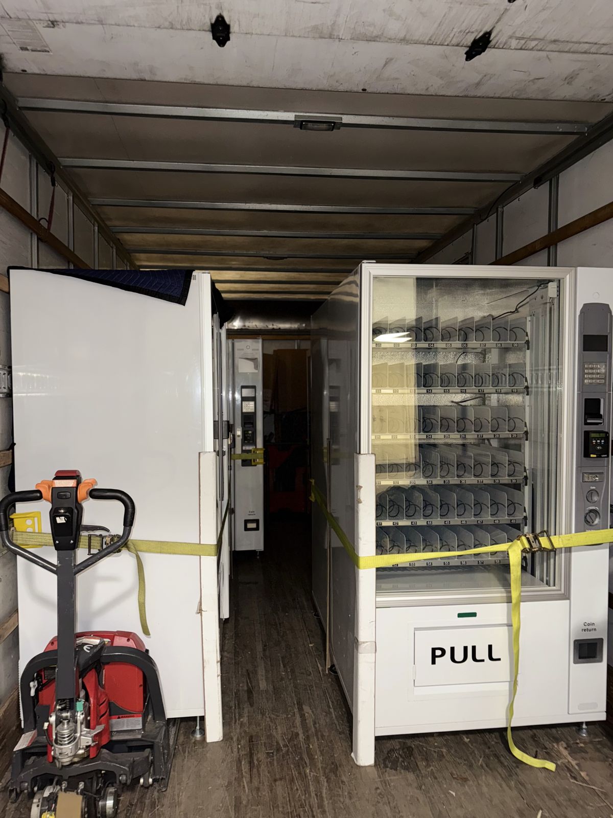 Two large vending machines are secured with TL Moving straps inside a truck beside a powered pallet jack.