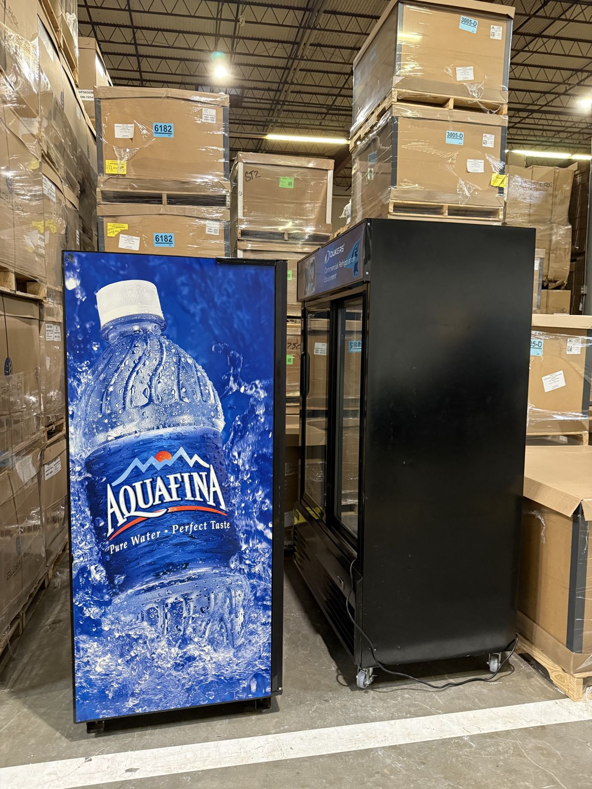 Two large beverage coolers stand among stacked pallets inside a warehouse.