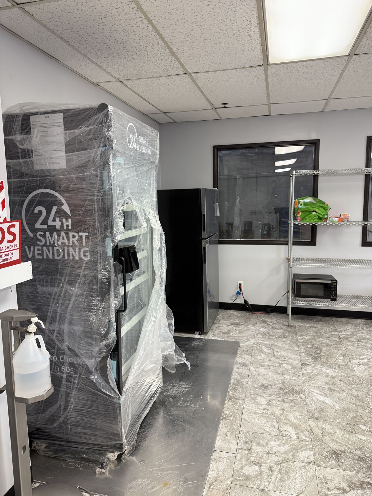 A commercial vending machine is wrapped for relocation beside a refrigerator and metal storage shelves.
